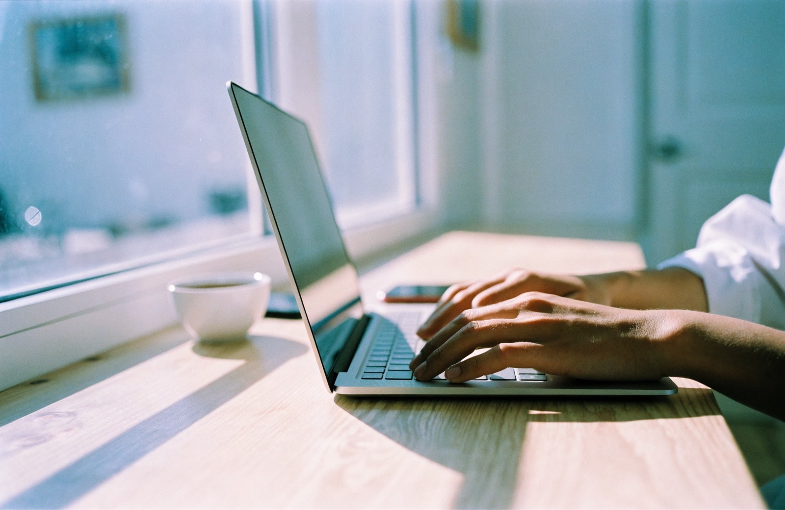 Hender på et laptoptastatur ved et lyst trebord i morgensol, med en kaffekopp ved siden av